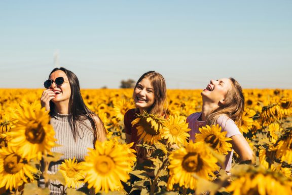 Drei Frauen lachen in einem Sonnenblumenfeld unter blauem Himmel.