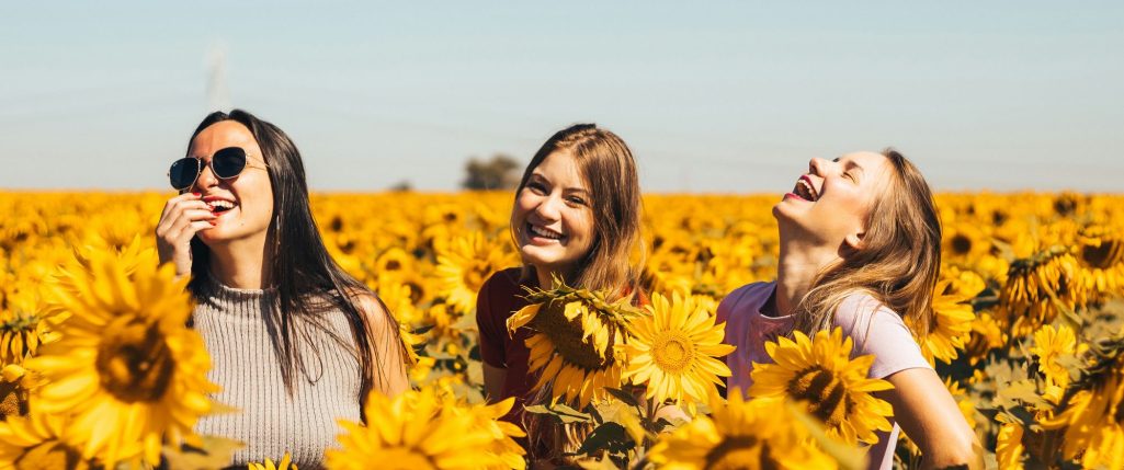 Drei Frauen lachen glücklich in einem Sonnenblumenfeld.
