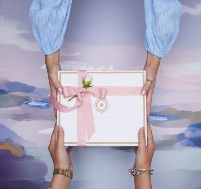 Zwei Hände übergeben ein Geschenk mit rosa Schleife und einer kleinen Blume.