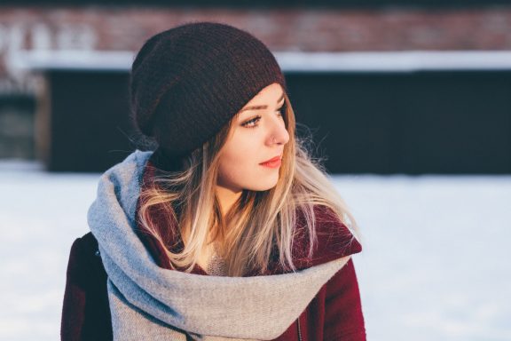 Junge Frau mit einem Schal und einer Mütze, steht im Schnee und schaut nachdenklich.