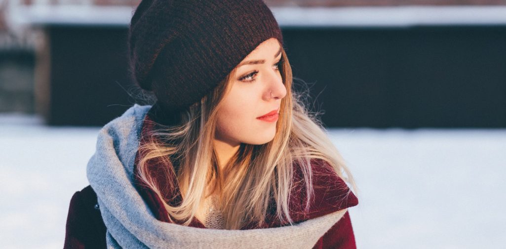 Junge Frau mit langen blonden Haaren, Hut und Schal in einer winterlichen Umgebung die überlegt welchen Weg sie einschlagen soll.
